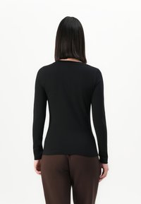 Persona con capelli neri lisci che indossa un top nero aderente a maniche lunghe e pantaloni marroni, girata di spalle su uno sfondo bianco.