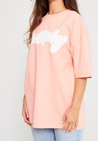 Světle růžové oversized tričko vyrobené z měkké bavlny, na přední straně se nachází výrazný bílý nápis "multiply", má krátké rukávy a kulatý výstřih.