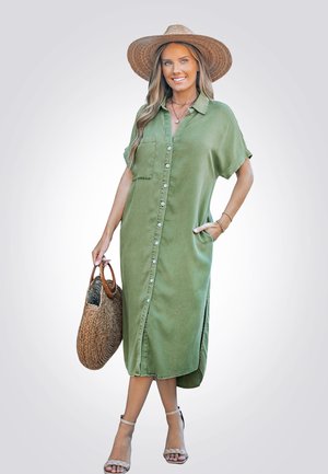 Olijfgroene blousejurk met korte mouwen, zijzakken en hoge splitten. Lichtgewicht stof met een relaxed fit, gecombineerd met een rieten handtas.