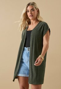 Grön, ribbad stickad kofta med korta ärmar och öppen front. Kombinerad med svart topp och ljusblå högmidjade shorts. Gyllene armringar synliga.