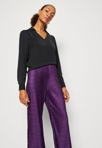 Blusa nera a maniche lunghe con scollo a V e una texture liscia, abbinata a pantaloni viola a vita alta e gamba larga con un motivo plissettato.