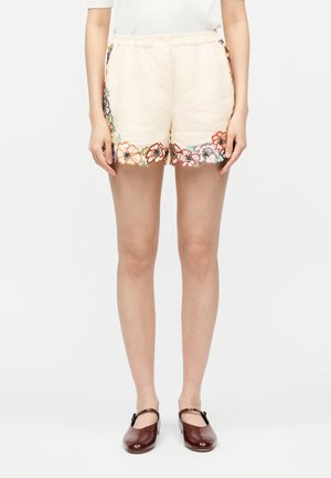 Γυναίκα που φοράει κρεμ σορτς με πολύχρωμη κεντητή λουλουδάτη διακόσμηση στις πλευρές και το τελείωμα, συνδυασμένα με σκούρα καφέ παπούτσια Mary Jane.
