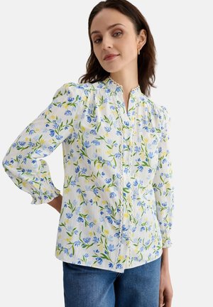 Mujer con blusa blanca con estampado floral azul y amarillo, tapeta de botones con borde ondulado, mangas largas abullonadas y vaqueros azules.