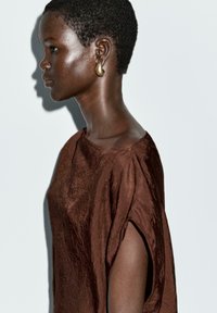 Hnědý, texturovaný oděv s širokými rukávy, nošený s velkým zlatým náušnicí, zobrazený v profilu na jednobarevném pozadí.