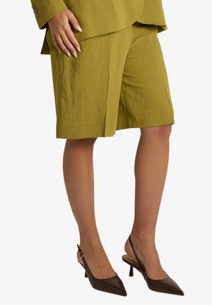 Donna che indossa shorts verde oliva fino al ginocchio, blazer abbinato e décolleté slingback marrone scuro con punta. La mano poggia sulla coscia.