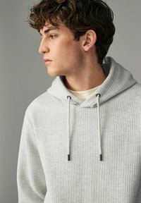 Sudadera gris acanalada con capucha ajustable, tejido texturizado y acentos metálicos negros en los cordones. Modelo mostrado de perfil.