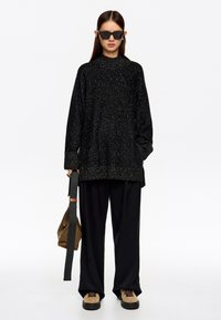 Suéter negro de gran tamaño con destellos metálicos, mangas largas y cuello alto, combinado con pantalones negros holgados y zapatos marrones. Bolsa marrón con correa.