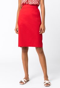 Punainen kynähame, jossa on istuva vyötärö, sileä rakenne ja polvipituinen muotoilu. Yhdistetty valkoisiin liukuviin sandaaleihin kontrastiksi.