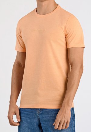 Mann trägt ein schlichtes pfirsichfarbenes T-Shirt mit kurzen Ärmeln mit dem Text "Lindbergh" und blaue Jeans vor einem einfarbigen hellen Hintergrund.