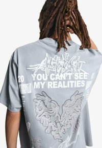 Grå kortærmet T-shirt med fede hvide tekster og indviklede linjetegninger på ryggen, med en løs og afslappet pasform samt blød stoftekstur.