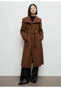 Bruine wollen blendjas met een grote kraag, een ceintuur in de taille en zakken, gecombineerd met zwarte wijde broek en glanzende schoenen.