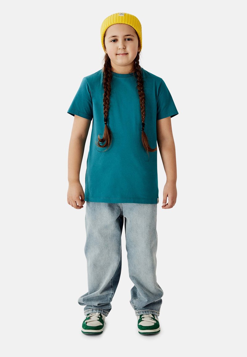 Turkis bomulds-t-shirt, løst lysblåt jeans, grønne og hvide sneakers, gul strikket hue. Langt brunt fletning-hår. Stående positur.