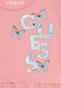 Ροζ βαμβακερό T-shirt με τη λέξη "GUESS" σε υφασμένα γράμματα, διακοσμημένο με γαλάζιες πεταλούδες και ασημένιες λεπτομέρειες. Λεπτομέρεια με βολάν στο τελείωμα.