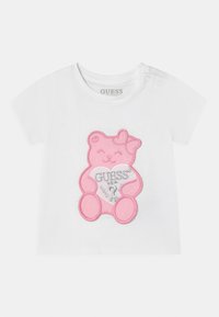 Tricou alb cu mânecă scurtă, având un grafic mare cu un urs roz care ține o inimă cu cuvântul "GUESS?" în argintiu. Două nasturi cu capse pe umăr.