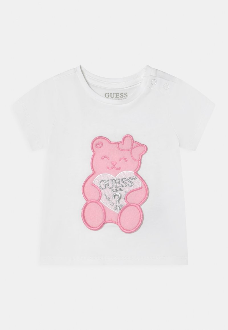 Tricou alb cu mânecă scurtă, având un grafic mare cu un urs roz care ține o inimă cu cuvântul "GUESS?" în argintiu. Două nasturi cu capse pe umăr.
