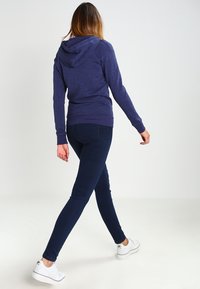 Marineblauwe hoodie met een trekkoord en gestructureerde stof, gecombineerd met donkerblauwe skinny jeans en witte sneakers, gezien vanaf de achterkant.
