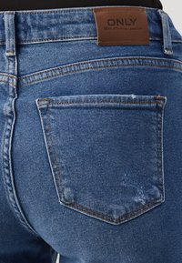 Plave traperice imaju stražnji džep s suptilnim istrošenim detaljem, kontrastnim narančastim šavom i smešim kožnim logotipom.