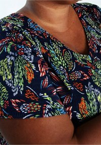 Gros plan sur une personne portant un haut bleu foncé avec un motif floral coloré, montrant le décolleté et la zone du haut du bras.