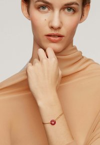 Pulsera de oro con un acento circular rojo, llevada en una mano de piel clara. El fondo presenta una textura de tela suave y neutra.