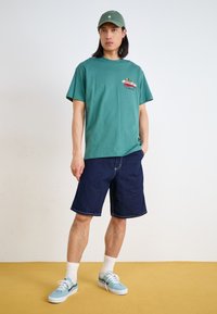 Camiseta de manga corta en color azul verdoso con un estampado gráfico, combinada con pantalones cortos de mezclilla azul oscuro y zapatillas azul claro con acentos blancos y calcetines.