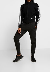 Schwarze Zip-Jacke mit weißen Streifen an den Ärmeln, kombiniert mit dunkelgrauen Jogginghosen. Beide aus weichem Stoff gefertigt und im entspannten Schnitt.