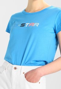 Modré bavlněné tričko s kulatým výstřihem a krátkými rukávy, s nápisem "G-STAR" v černé, růžové a světle modré barvě napříč hrudí.