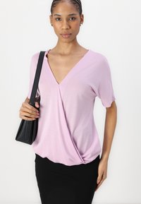 Haut court lavande à manches courtes avec un décolleté en V et un détail de ourlet torsadé, associé à une jupe noire ajustée et un sac bandoulière noir.