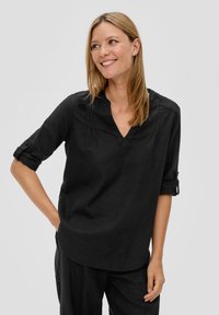 Blouse en lin noir avec un col en V, manches 3/4 et poignets retroussés ; présente de subtils plis près du col ; texture lisse.