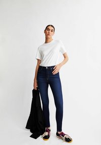Weißes T-Shirt mit kurzen Ärmeln, hoch taillierte dunkle Skinny-Jeans aus Denim und schwarze Sneakers mit pinken Schnürsenkeln, hält eine schwarze Jacke.