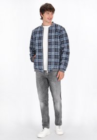 Navy plaid siuvinėtas švarkas su pilkais akcentais, su užtrauktuku priekyje ir šoninėmis kišenėmis, dėvimas ant baltos marškinėlių ir pilkų džinsų. Balti sportbačiai.