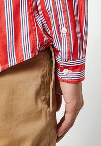 Adulto che indossa una maglietta a maniche lunghe a strisce rosse, bianche e blu con polsini abbottonati e pantaloni marrone chiaro, con la mano appoggiata accanto alla tasca.