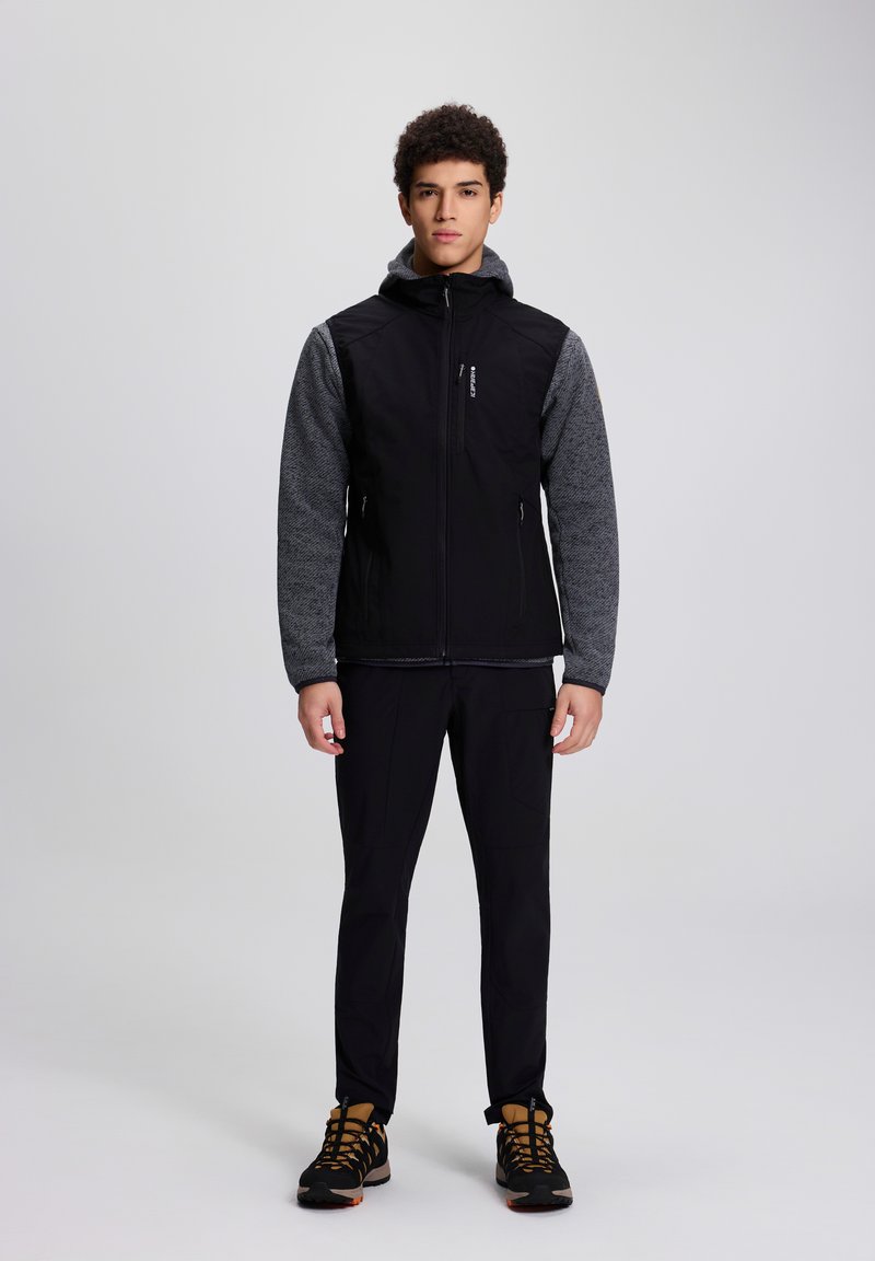Junger Mann steht, trägt schwarze Zip-up-Jacke mit grauen Ärmeln, schwarze Hose und braun-schwarze Wanderschuhe vor schlichtem hellem Hintergrund.