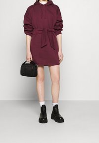 Bordeauxfarbenes Hoodie-Kleid mit einem Gürtel, im lässigen Schnitt, mit übergroßen Ärmeln und einer weichen Textur. Kombiniert mit klobigen schwarzen Stiefeletten und einer kleinen Tasche.