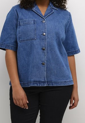 Person trägt ein kurzärmliges blaues Jeanshemd mit vorderen Knöpfen und Tasche, kombiniert mit einer schwarzen Hose vor einfarbigem Hintergrund.