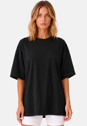 Femme aux cheveux blonds mi-longs portant un t-shirt noir ample à manches courtes et un short blanc, debout devant un fond blanc.