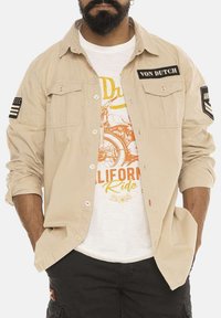 Chemise beige à boutons avec deux poches poitrine, ornée de patches "VON DUTCH" et de drapeaux sur les manches. Portée sur un T-shirt graphique blanc.