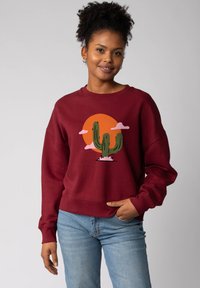 Bordeaux sweatshirt met een ontspannen pasvorm met een cactusontwerp tegen een oranje zon en roze wolken. Gemaakt van een katoenmix met ribgebreide manchetten.