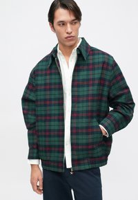 Πράσινο καρό μπουφάν με κόκκινες λεπτομέρειες, με φερμουάρ μπροστά, υπερμεγέθες μέγεθος και πλαϊνές τσέπες, φορεμένο πάνω από ένα λευκό πουκάμισο. Υφή υφάσματος.