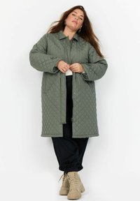 Cappotto lungo trapuntato di colore verde oliva con chiusura a bottoni, tasche grandi e colletto. Abbinato a pantaloni neri e sneakers beige.