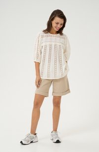 Blusa blanca de eyelet con mangas acampanadas, combinada con pantalones cortos de pana color beige. Modelo con zapatillas deportivas blancas, de pie contra un fondo liso.