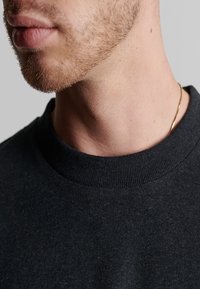 Sort crewneck sweatshirt med en ribbet krave, der har et glat, tekstureret stof. Subtle syninger og en tynd guldkæde er synlige.
