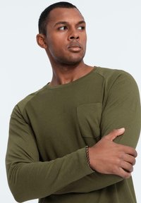 Camisa de manga larga verde oliva con textura acanalada, cuello redondo y bolsillo en el pecho; el modelo cruza los brazos, mostrando el ajuste de la camisa.