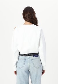 Witte sweatshirt met lange mouwen en uitgesneden schouders, gecombineerd met lichtblauwe denim jeans met een zwart merkembleem in de taille.