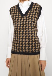 Neuleliivi, jossa on musta ja ruskea houndstooth-kuvio, V-kaula-aukko, hihaton malli ja ribattu helma. Yllä valkoinen paita.