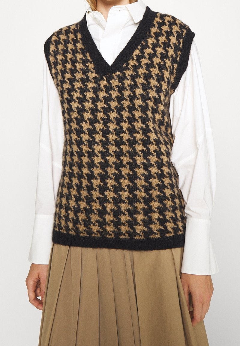 Neuleliivi, jossa on musta ja ruskea houndstooth-kuvio, V-kaula-aukko, hihaton malli ja ribattu helma. Yllä valkoinen paita.