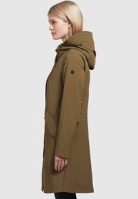 Parka olive green, mi-longue, avec une capuche, des poches latérales et une texture lisse. Comprend des cordons ajustables et un patch logo discret.