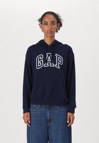 Marinblå hoodie med ordet "GAP" i vit text, med en framficka, dragsko i huvan, samt ribbat nederkant och ärmslut. Avslappnad passform.