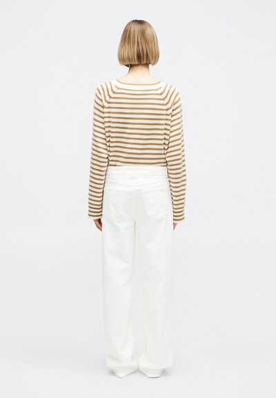 Femme aux cheveux blonds courts debout, portant une chemise à manches longues beige à rayures blanches et un pantalon blanc ample, sur un fond uni.