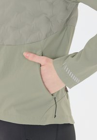 Ljusgrön jacka med en texturerad övre del, en sidoficka och reflektiva accentband på ärmarna. Lättviktsmaterial.