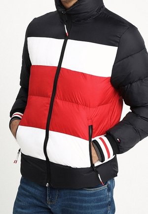 Mann trägt eine farbige Pufferjacke mit schwarz, weiß und rot horizontalen Streifen, die Hände in den Reißverschlusstaschen, kombiniert mit blauen Jeans.